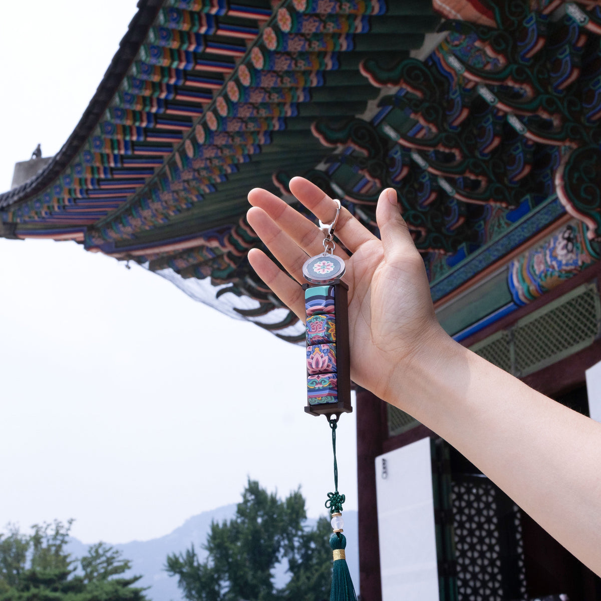 Hands holding a Black Dancheong keycap keychain in front of a Korean palace building